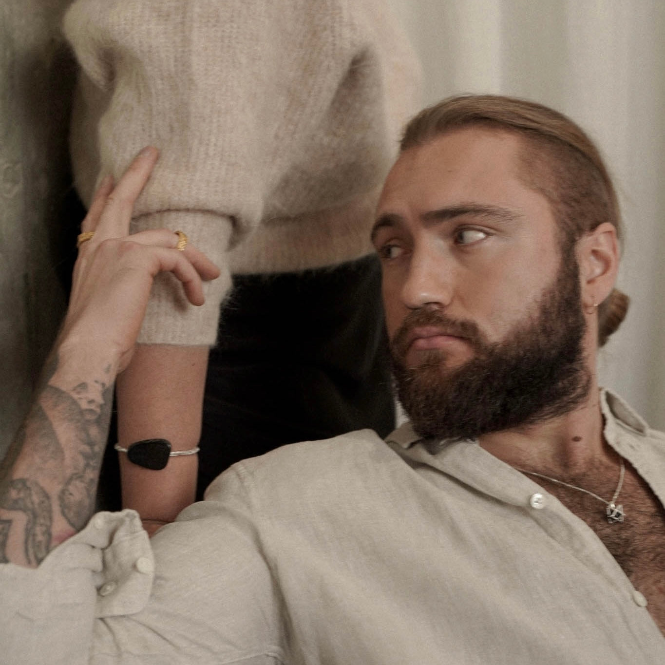 Man with a beard wearing a light-colored shirt, rings and necklace and touching beautiful woman's hand with statement handmade jewellery wearing a beige sweater